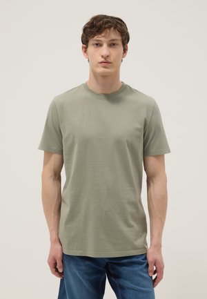 Young man with short brown hair wearing olive green short-sleeve t-shirt and blue jeans, standing against a plain light background.