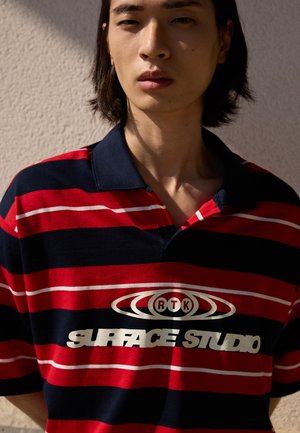 Giovane con capelli alla lunghezza delle spalle che indossa una polo a righe rosse e blu navy con il logo "RTK Surface Studio", in piedi contro un muro testurizzato.