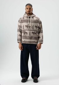 Beige hoodie met patroon en zwarte paarden- en cactussymbolen, voorzien van een capuchon met trekkoord en brede mouwen, gecombineerd met donkerblauwe, losse broek.