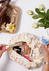 Pochette cosmetica in tessuto crema con motivi colorati di gelato e dolci, riempita con vari prodotti di trucco e una tazza di caffè nei dintorni.