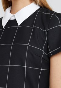 Svart klänning med vit krage, prydd med ett rutmönster i vita linjer. Strukturerad tyg med korta ärmar som betonar det geometriska designen.