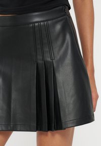 Jupe en cuir plissée noire portée avec un top noir, montrant une longueur mi-cuisse et un détail de fermeture éclair sur le côté.