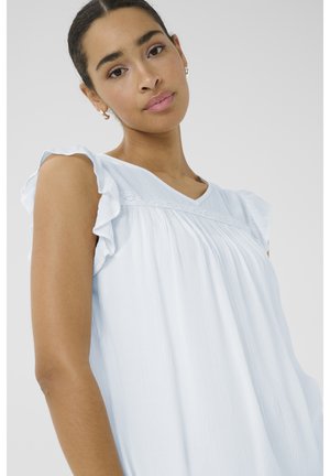 Ung kvinde med mørkt hår bundet tilbage iført en lyseblå ærmeløs top med flæser på skuldrene, der ser blidt mod kameraet.