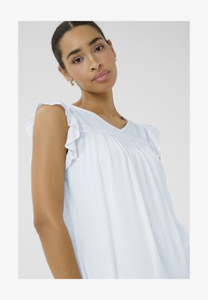 Ung kvinde med mørkt hår bundet tilbage iført en lyseblå ærmeløs top med flæser på skuldrene, der ser blidt mod kameraet.