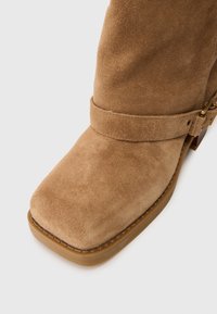 Stivale alla caviglia in suede marrone chiaro con punta quadrata, dettaglio di fibbia e cinturino, e suola in gomma spessa su sfondo bianco.
