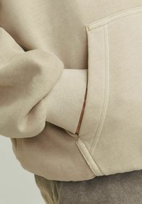 Beige mikina s ribovanými manžetami, boční kapsou a měkkou texturou. Detailní pohled na rukáv a švy.