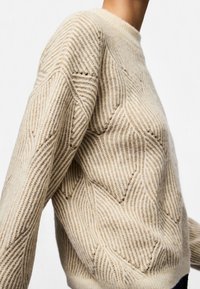 Suéter de punto beige con patrones geométricos texturizados, llevado por una persona, mostrado desde el hombro hasta la cintura sobre un fondo blanco.