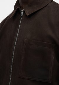 Ruskea mokkatakki terävällä kauluksella, hopeanvärisellä vetoketjulla ja suurella rintataskulla. Sileä pinta ja yksinkertainen muotoilu.