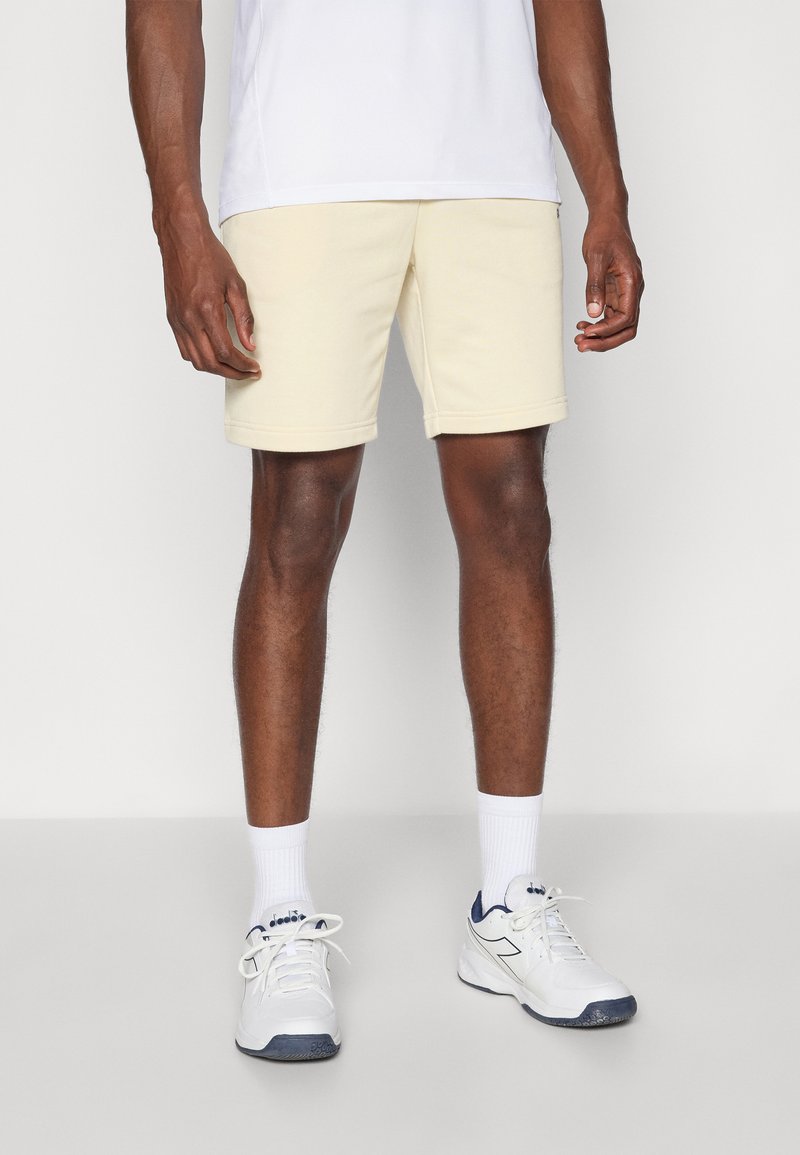 Cremfarbene Shorts aus einem weichen Stoff mit elastischem Bund, kombiniert mit weißen Sneakers und gerippten weißen Socken.