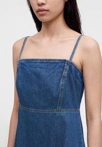 Jeune femme portant une robe en jean bleue sans manches avec de fines bretelles, visible des épaules au milieu du torse sur un fond uni.