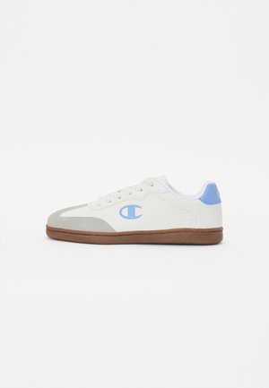 Zapatilla blanca con puntera gris claro, logotipo azul en el lateral, parche azul en el talón, cordones blancos y suela marrón, mostrada desde el lado exterior.