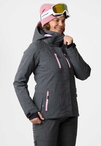 Dunkelgraue wasserdichte Jacke mit pinken Reißverschlüssen, ausgestattet mit einem hohen Kragen, einer Kapuze und einem strukturierten Stoff. Getragen mit Skibrille und einer pinken Mütze.