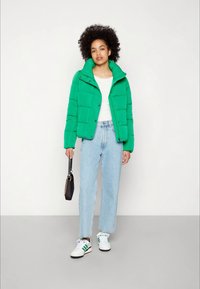 Green puffer jacket with a high collar, zip closure, and horizontal stitching; paired with light blue jeans and white sneakers.