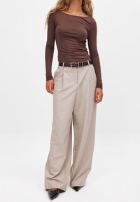 Ruskea pitkähiha toppi yhdistettynä korkeavyötäröisiin, leveälahkeisiin beigeihin housuihin, joissa on ruskea vyö; mustat kärkitossut täydentävät asukokonaisuutta.