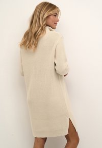 Stickad klänning i beige med hög krage, ärmar i medellängd, sidofickor och ribbade muddar och fåll. Strukturerad yta.