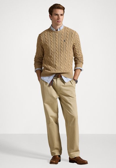 Beige kabelgebreide trui over een lichtblauw gestreept shirt, gecombineerd met kaki broek en bruine instappers. Eenvoudig, textuur ontwerp zonder zichtbare logo's.