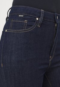 Vaqueros de mezclilla azul oscuro con una textura suave, costuras amarillas, acentos metálicos y una etiqueta de marca visible en la cinturilla.