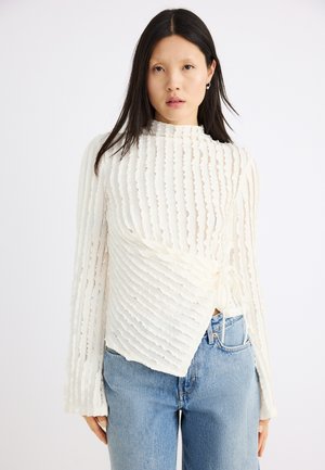 Kvinde med langt mørkt hår iført en tekstureret hvid langærmet top med omslagsdesign og lyseblå jeans, stående mod en hvid baggrund.