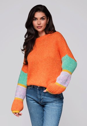 Pomarańczowy sweter robiony na drutach z kolorowymi wzorzystymi rękawami w miętowej zieleni, purpurze i żółci. W połączeniu z niebieskimi dżinsami.