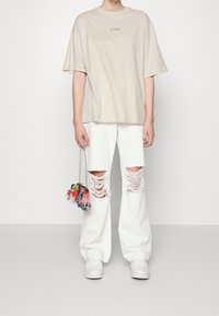 Camiseta oversized beige con el texto "VENUS", combinada con jeans anchos blancos desgastados. Zapatillas blancas y un bolso de flecos colorido completan el look.
