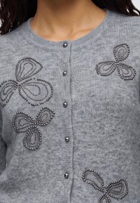 Pull gris à boutons avec des grappes décoratives de perles noires formant des formes abstraites de papillons sur le devant.