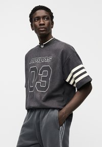 Γκρι κοντομάνικη μπλούζα από κορντόν με λευκές ρίγες και το «ADIDAS 03» τυπωμένο μπροστά. Το μοντέλο φοράει γκρι φούτερ παντελόνι με κάθετες ρίγες.