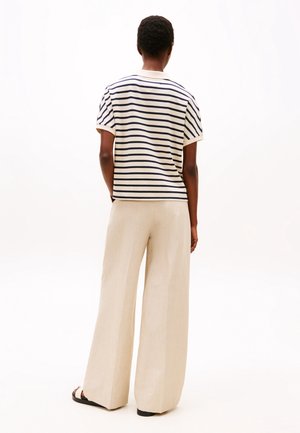 Person, der står med ryggen til iført en hvid kortærmet skjorte med marineblå striber, beige bukser med vide ben og sorte sandaler.