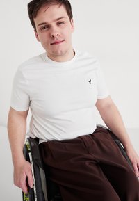 Λευκό βαμβακερό T-shirt με μικρό μαύρο λογότυπο στο στήθος, συνδυασμένο με καφέ παντελόνι, φορεμένο από ένα άτομο που κάθεται σε αναπηρικό καρότσι.