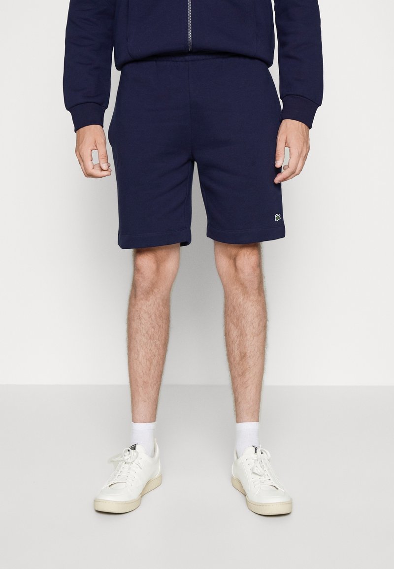 Personne portant un short bleu marine, des chaussettes blanches et des baskets blanches, debout les mains relâchées le long du corps sur un fond uni.