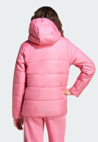 Eine Person mit lockigem Haar trägt eine leuchtend pinkfarbene gepolsterte Jacke mit Kapuze, von hinten gesehen, und steht mit einer Hand auf der Hüfte.
