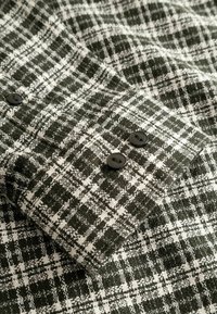 Tissu à carreaux texturé noir et blanc avec deux boutons noirs sur un poignet plié, montrant un motif tissé détaillé et des coutures.