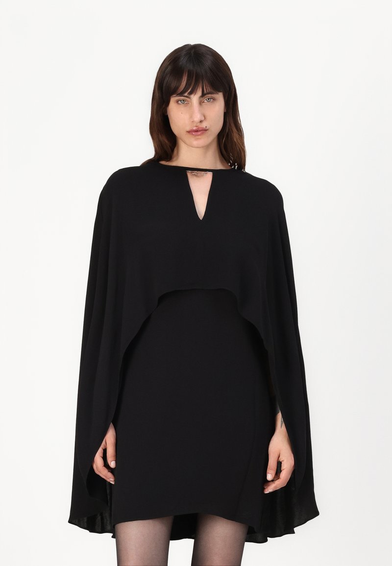 Črna obleka s plaščem, ki ima V-izrez, tekočo tkanino in slojevito zasnovo, ki sega do komolcev, kar ustvarja dramatično silhueto.