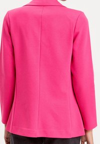 Blazer rose avec une texture lisse, des épaules structurées et des manches longues. Comprend une fente unique à l'arrière et des détails de couture soignés.