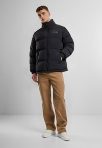Jachetă puffer neagră cu guler înalt și detalii cusute, având două buzunare laterale și manșete elastice, purtată peste pantaloni bej și pantofi albi.