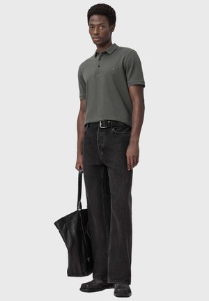 Man draagt een donkergroen poloshirt, zwarte riem, zwarte wijde jeans en zwarte schoenen, en houdt een grote zwarte leren draagtas vast.