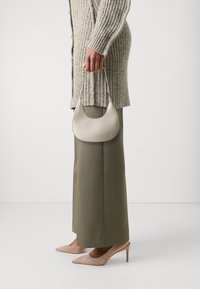 Sac à main en cuir gris clair en forme de croissant, porté par une personne vêtue d'un pull côtelé beige et d'un pantalon vert olive, assorti à des talons beige nude.