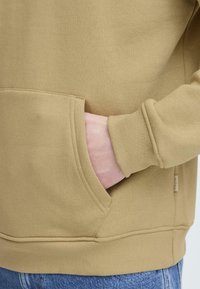 Khaki hoodie med en kengurulomme, ribbede manchetter og kant, lavet af blødt stof med en glat tekstur, båret sammen med blå jeans.