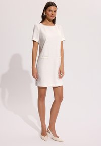 Kobieta ubrana w białą, teksturowaną sukienkę z krótkim rękawem i przednimi kieszeniami, w połączeniu z białymi, wąskimi czółenkami na obcasie na pasku oraz srebrnymi kolczykami.