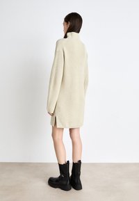Abito lavorato a maglia di colore crema con colletto alto, maniche lunghe, fessure laterali e vestibilità rilassata, abbinato a stivali neri fino alla caviglia.