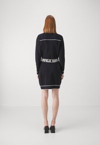 Eine Frau mit glattem roten Haar steht mit dem Rücken zu uns und trägt ein schwarzes Kleid mit weißem Saum und einem gebrandeten Gürtel, kombiniert mit schwarzen High Heels.