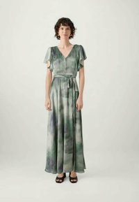 Γυναίκα με κοντά σγουρά μαλλιά φοράει ένα μέγιστο φόρεμα tie-dye σε πράσινο και γκρι χρώμα με φτερούγες και ζώνη στη μέση, συνδυασμένο με μαύρες ανοιχτές μποτες.