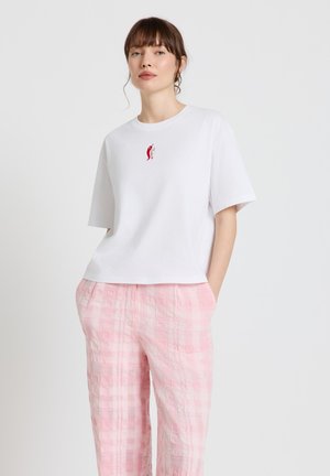 Femme aux cheveux bruns portant un t-shirt blanc avec un logo piment rouge et un pantalon ample à carreaux roses, debout les mains dans les poches.