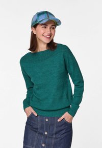 Grüner Strickpullover mit einem Bootsausschnitt, kombiniert mit einem Jeansrock mit Knopfleiste. Die Mütze hat ein blaues Karomuster.