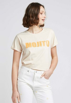 T-shirt beige avec "MOJITO" en orange vif, à manches courtes et col rond, associé à un pantalon blanc avec de la broderie florale.