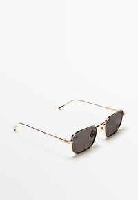 Zonnebril van goudkleurig metaal met vierkante donkere glazen, voorzien van slanke pootjes en subtiele scharnierdetails, geplaatst tegen een witte achtergrond.