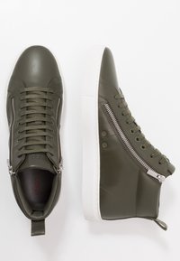 Olijfgroene high-top sneakers van textured leer, met witte rubberen zolen, veters en zilveren ritsen aan de zijkanten voor extra detail.