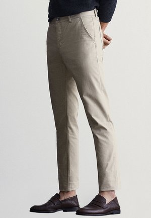 Personne portant un pantalon beige cintré et des mocassins en cuir marron foncé, debout devant un fond clair uni.