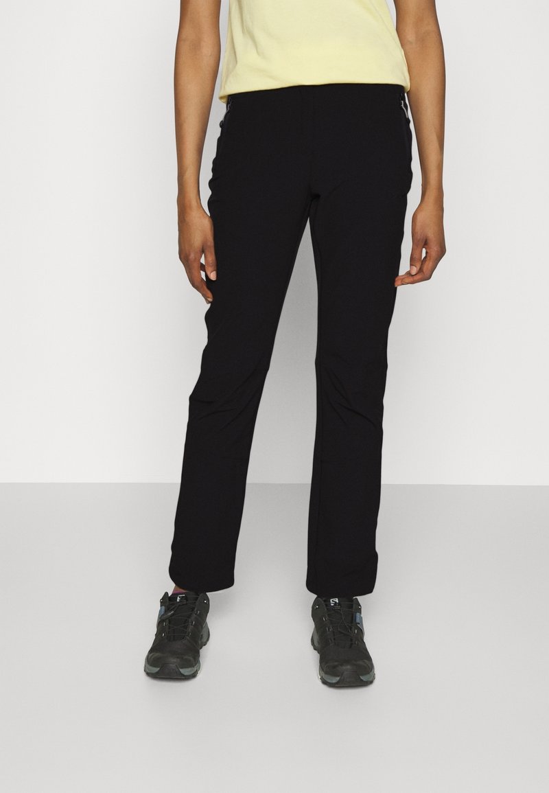 Personne portant un pantalon droit noir, des chaussures de randonnée noires, et un haut jaune clair à manches courtes sur un fond clair uni.