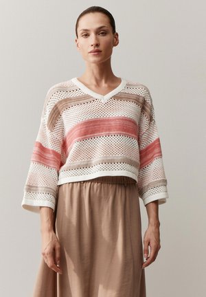 Frau trägt einen locker gestrickten Pullover mit V-Ausschnitt, rosa und beige horizontal gestreift, und einen hoch taillierten beigen Rock, steht vor einer schlichten grauen Wand.