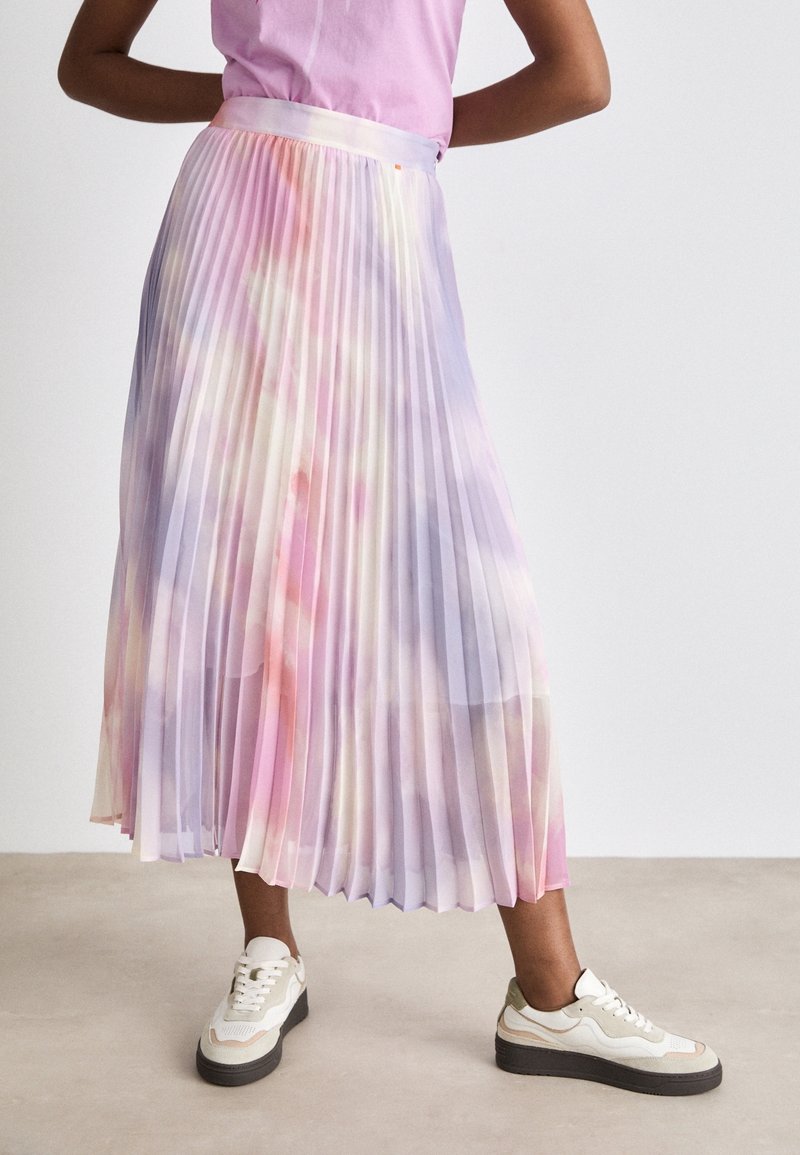 Femme portant une jupe midi plissée tie-dye pastel assortie à des baskets blanches et beiges, debout les mains sur les hanches.